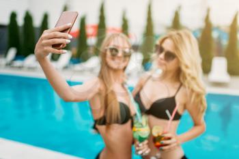 Two sexy girls in swimsuits and sunglasses makes selfie near the swimmimg pool. Slim women sitting by the poolside, resort holidays 