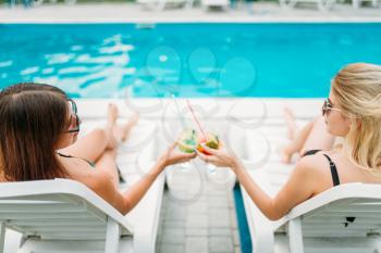 Two sexy girls relax with cocktails on deck chairs near the swimmimg pool. Slim women sitting by the poolside, resort holidays