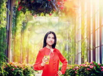 Cheerful woman in shirt and straw hat drinks freshly squeezed orange juice, park background. Young girl with yellow vitamin beverage, healthy lifestyle