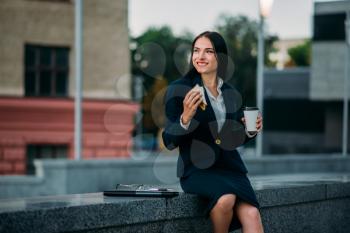 Happy business woman with sandwich and coffee in hands, lunch outdoor. Modern building, financial center, cityscape. Female businessperson in suit at workplace
