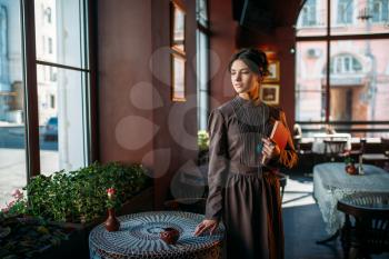 Young female person stands inside of cafe looking in window. Retro style lady in vintage cafe with red book in her hands. Dark hair young woman in vintage dress and jewelry.