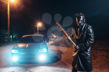 Serial maniac in leather coat and hockey mask, bloody baseball bat wrapped in metal chain in hands, against black car with light at the night. Horror, bloody murderer, murder weapon