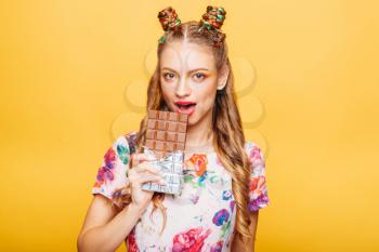 Beautiful young woman with playful look bites huge chocolate. Stylish girl with blonde curly hair. Stylish girl in colorful summer dress, yellow wall on background.
