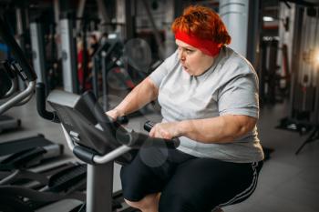 Fat woman training on exercise bike in gym. Calories burning, obese female person in sport club