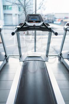 Treadmill against big window, gym interior, nobody, jogging track, stationary running simulator, sport equipment in fitness club