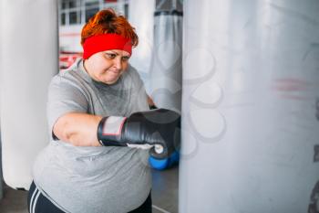 Fat woman in boxing gloves works with punching bag, workout in gym. Calories burning, obese female person on hard training in sport club