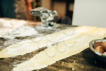Pasta machine, dough and eggs on wooden kitchen table sprinkled with flour, nobody. Traditional italian cuisine