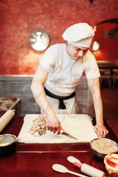 Male chef prepares apple strudel on wooden kitchen table, pastry ingredients on background. Homemade sweet dessert, pie preparation process