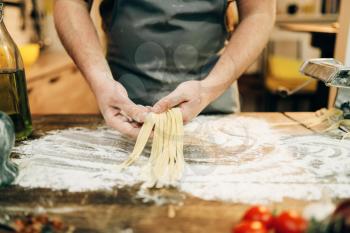 Male chef in apron cooking dough and prepares pasta machine on wooden kitchen table