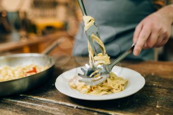 Male chef puts fresh pasta in a plate, pan on wooden kitchen table. Homemade fettuccine preparation process. Traditional italian cuisine