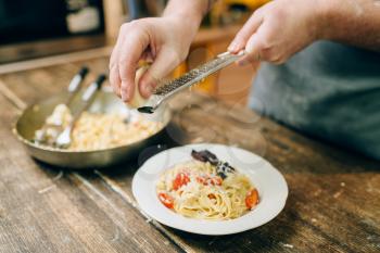 Male chef hands grates cheese in to the plate with fresh cooked fettuccine, pan on wooden kitchen table. Homemade pasta preparation process. Traditional italian cuisine