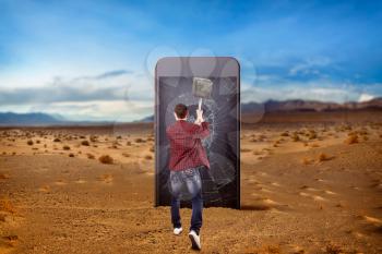 Phone addicted people, loneliness in the net concept. Man smashes a large smartphone screen with a sledgehammer, desert valley on background. Manipulation of consciousness. Scaling effect