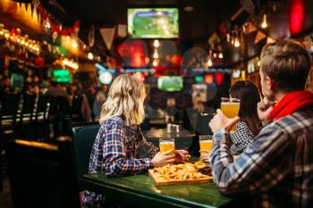 Football fans watching match and drinks beer at the table in sports bar. Tv broadcasting, young friends leisures in pub