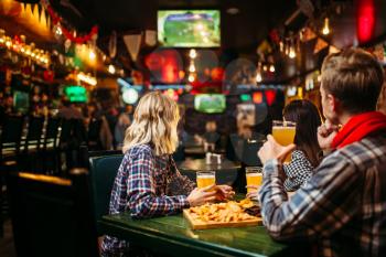 Football fans watching match and drinks beer at the table in sports bar. Tv broadcasting, young friends leisures in pub