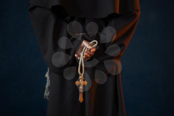 Male monk in black robe holds wooden rosary and cross in hands, religion. Mysterious friar in dark cape