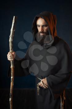 Medieval monk in black robe with hood rests on a stick, religion. Mysterious friar in dark cape
