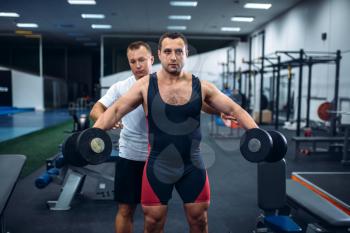 Male weightlifter doing exercise with dumbbell under trainer control, gym interior on background. Weightlifting workout in sport or fitness club