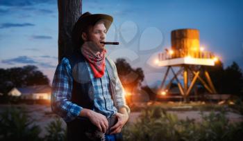Brutal cowboy smokes a cigar on sunset, evening relax on texas ranch, western. Vintage male person with gun on farm, wild west culture