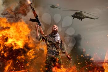 Bearded terrorist with rifle in hands stands in explosion and fire. Terrorism and terror, soldier in khaki camouflage, military helicopters in the sky on background