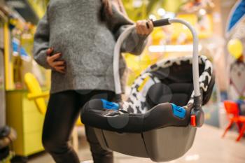Pregnant woman with portable bed choosing child car seat in store. Goods for safe transportation of children