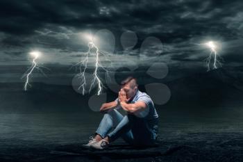 Young man sitting on the ground in desert, storm with lightning flash on background