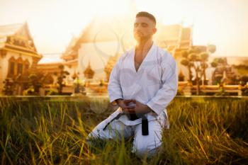 Male karate fighter sitting on the ground against ancient temple. Martial art training outdoor on sunset. Photo manipulation with background