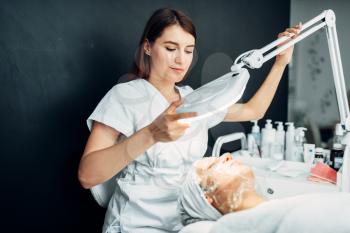 Beautician watches on patient face in mask, getting rid of wrinkles, cosmetology clinic. Facial skincare, rejuvenation procedure in spa salon, health care