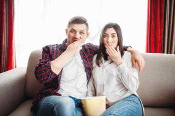 Smiling couple sitting on couch and watch tv at home, woman with remote control in hand, window on background