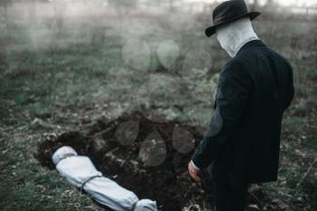 Bloody murderer stands against a grave with victim in forest, the body wrapped in a canvas, serial maniac concept