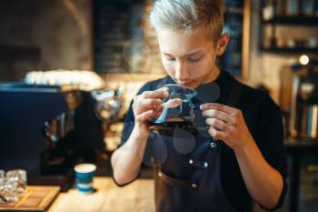 Young male barista sniffs fresh prepared black coffee, cafe counter on background. Professional espresso preparation by barman in cafeteria, bartender occupation