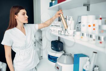 Young female beautician with bottle of cream in hands, cosmetology cabinet. Doctor in white uniform in spa salon, beauty medicine