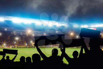 Group of football fans on tribune are cheering for their team victory, back view, crowd of people silhouettes with hands up holds slogans and banners on sport arena 