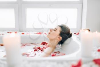 Young woman sleeps in the bath with foam, rose petals and burning candles decor. Full relaxation, romantic setting in bathroom