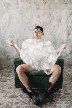 Funny freak man weared in swimming goggles sitting on a chair, grunge room interior. Nerd in abandoned house