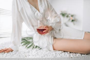 Female person in white bathrobe sitting on the edge of the bath with glass of red wine. Bathroom interior with window on background