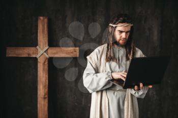 Man in the image of Jesus Christ uses laptop, crucifixion cross on black background. Believers and modern technologies concept