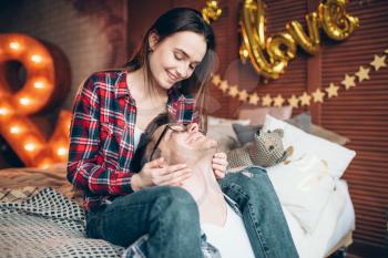 Young wife and husband hugs on the bed in bedroom with lighting decoration. Love couple in clothes embrace on couch