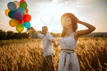 Love couple with balloons, leisure in a rye field on sunset. Pretty wife and husband walking on summer meadow