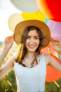 Cute woman in white dress and straw hat holds bunch of colorful balloons. Pretty woman on summer meadow at sunny day