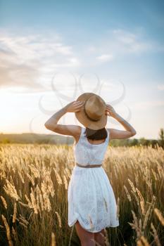 Woman in white dress and straw hat walking in wheat field on sunset. Female person on summer meadow