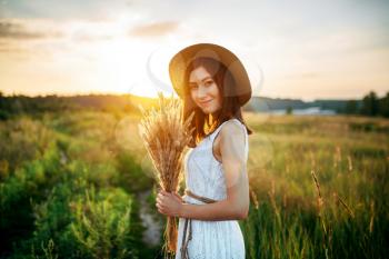 Cute woman in white dress and straw hat holds wheat bouquet in the field at sunset. Pretty girl on green summer meadow