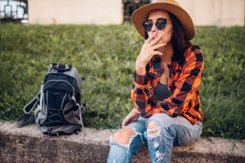 Female tourist in hat and sunglasses smoke cigarettes, break during the tour. Summer adventure of young woman, city walking