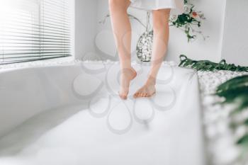 Female person legs dips into the bath with foam. Bathroom interior with window on background