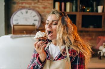 Housewife in apron eating sweet dessert with cream, kitchen interior on background. Female cook tastes fresh homemade cake. Chef eats pie