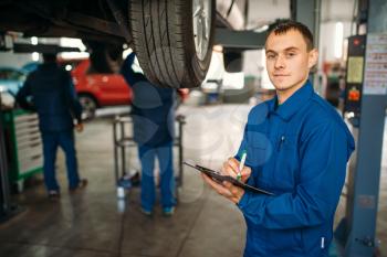 Repairman with notebook fills the check list, car on the lift, fixing the problems, suspension diagnostic. Automobile service, vehicle maintenance