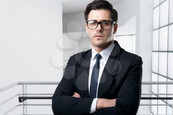 Businessman in glasses, tie and black suit poses in modern business center with glass walls