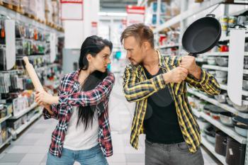 Young couple plays with frying pan and rolling pin in supermarket. Male and female customers on family shopping. Man and woman purchasing goods for the house
