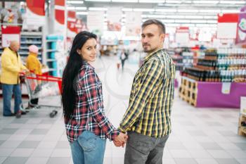 Young couple hold hands in supermarket, back view. Male and female customers on family shopping. Man and woman purchasing in hypermarket