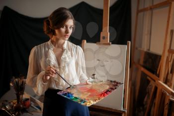 Female painter with color palette and brush standing against easel in studio. Creative paint, woman drawing pencil sketch