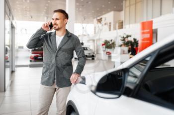 Man talking by phone near the new car in showroom. Male customer buying vehicle in dealership, automobile sale, auto purchase